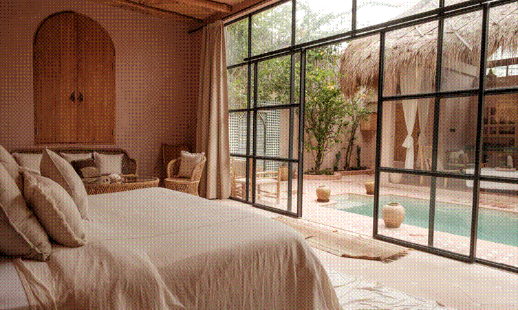 A sunlit, earth-toned bedroom with a bed dressed in beige linens and rattan seating, opening through large black‑framed glass doors onto a rustic courtyard featuring a small turquoise pool, terracotta pots, and a thatched‑roof structure.